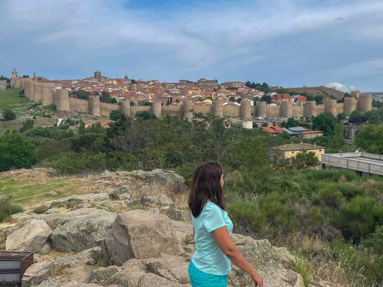 Una viajera de espaldas contempla la impresionante muralla medieval de Ávila y el casco histórico desde el mirador de Los Cuatro Postes, bajo un cielo nublado.