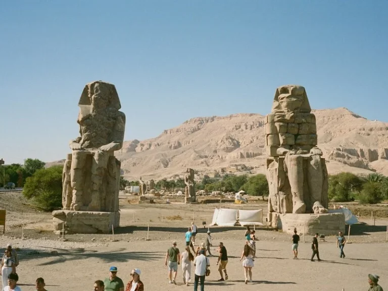 Dos estatuas colosales de piedra sentadas sobre el terreno árido de Luxor; la presencia de visitantes en la base resalta la magnitud y altura de las esculturas frente al horizonte.