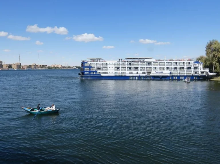 Una embarcación navegando por las tranquilas aguas del río Nilo, con palmeras y vegetación densa en la orilla.