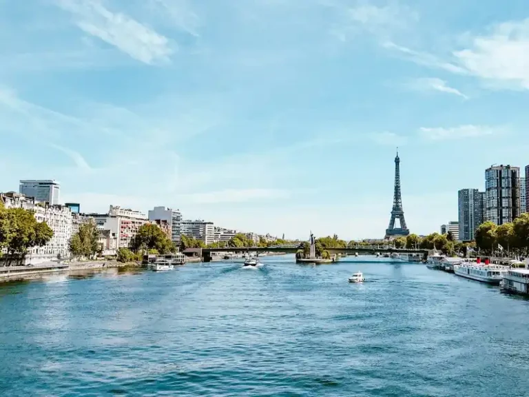 Fotografía panorámica del río Sena en París bajo un cielo azul, con barcos navegando y la Torre Eiffel al fondo, flanqueada por edificios de la ciudad.
