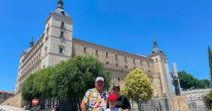 una imagen con una familia de madre, padre e hijo, con el alcazar de toledo de fondo, con un cielo azul vibrante.