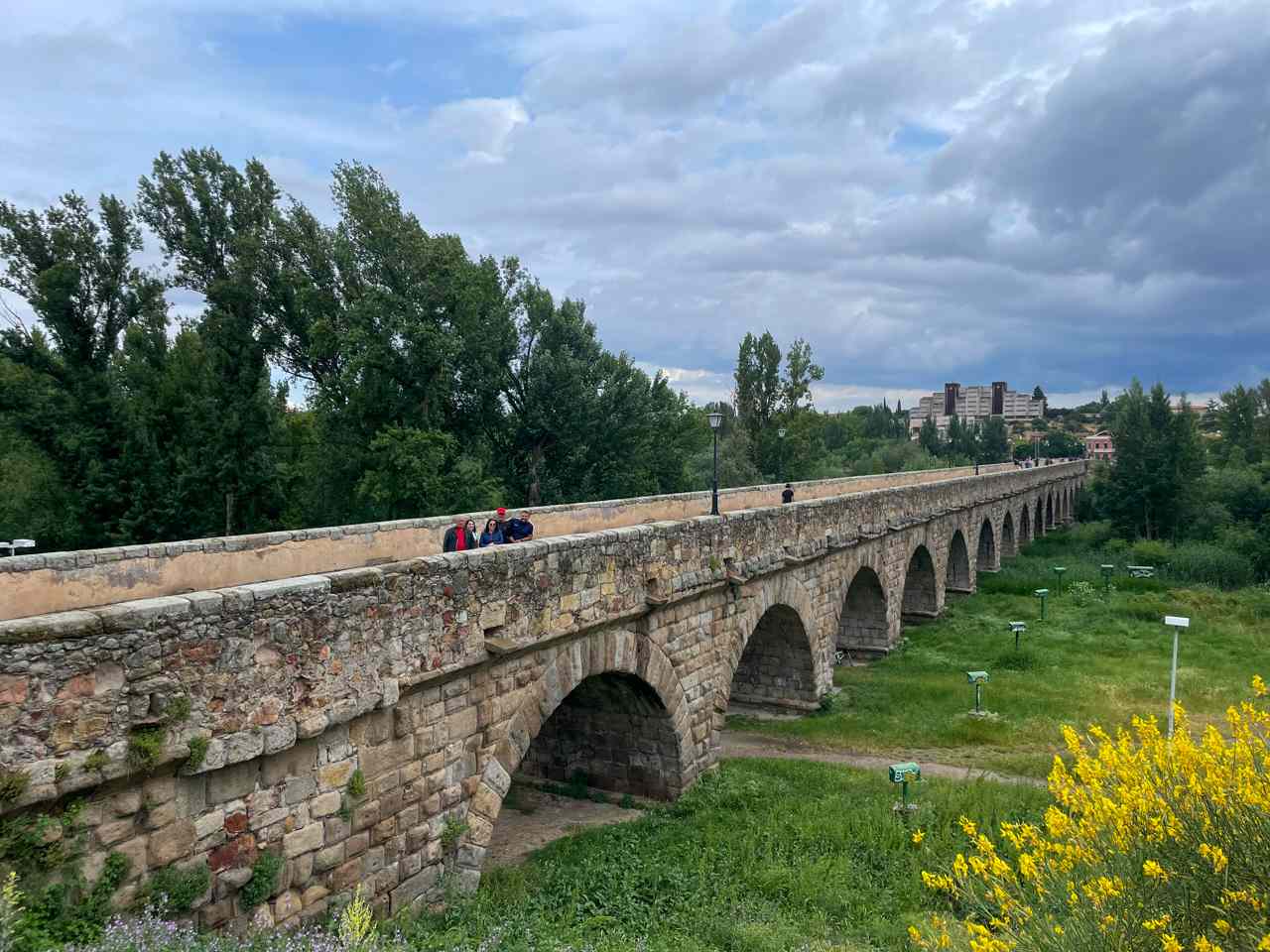 Vista panorámica del histórico Puente Romano de Salamanca con sus múltiples arcos de piedra. Un grupo de cuatro viajeros de G-travel está de pie sobre el puente. El fondo muestra árboles verdes y edificios de la ciudad bajo un cielo nublado.